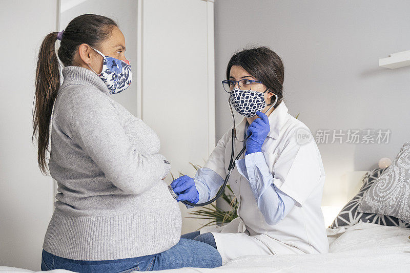 Home caregiver. Doctor's visit to a young pregnant woman at home during COVID-19 pandemic. Both wearing protective face masks for illness prevention. Helping hand during the quarantine to flatten the curve.